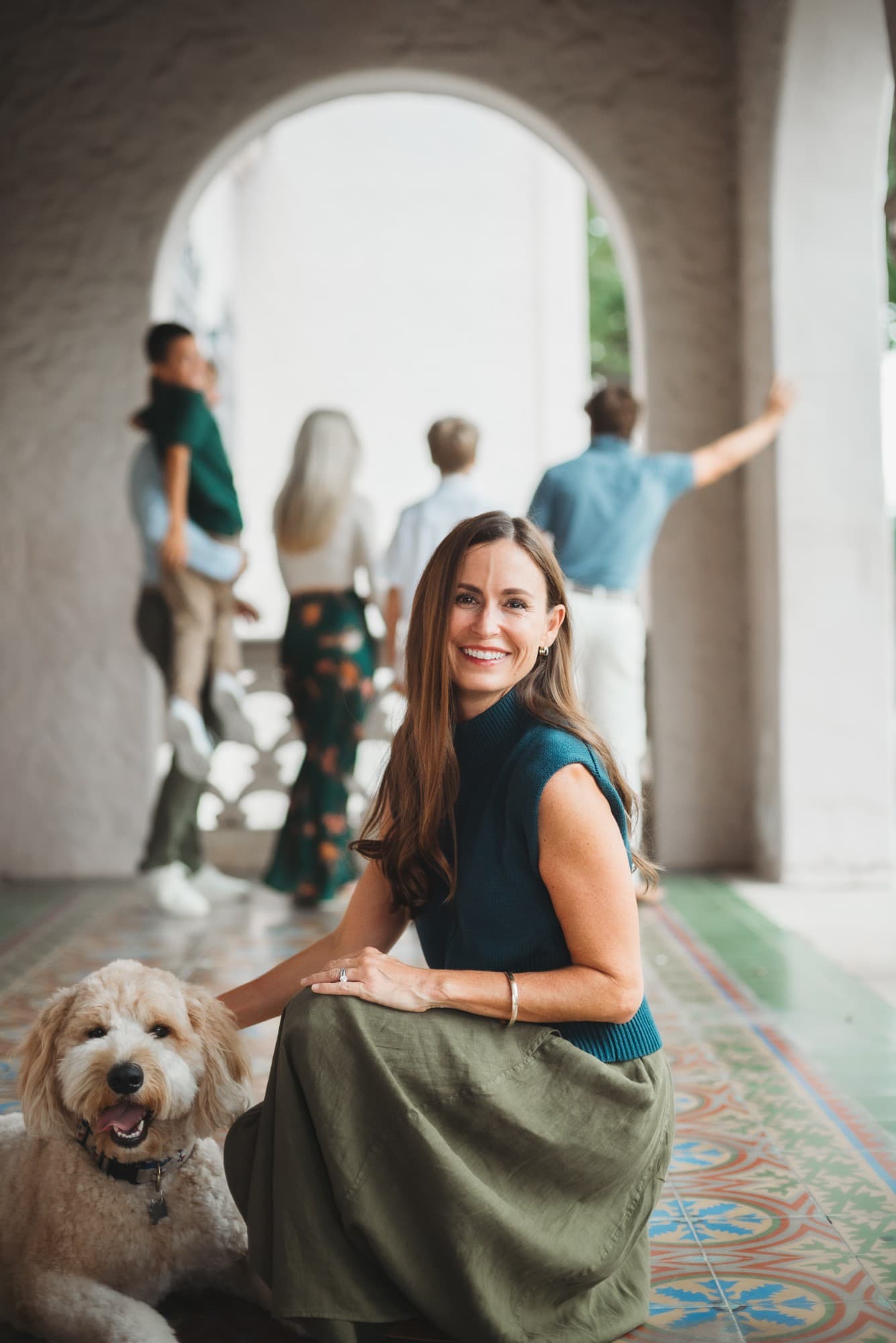 Dominique kneeling beside her goldendoodle, with her family under an archway behind her
