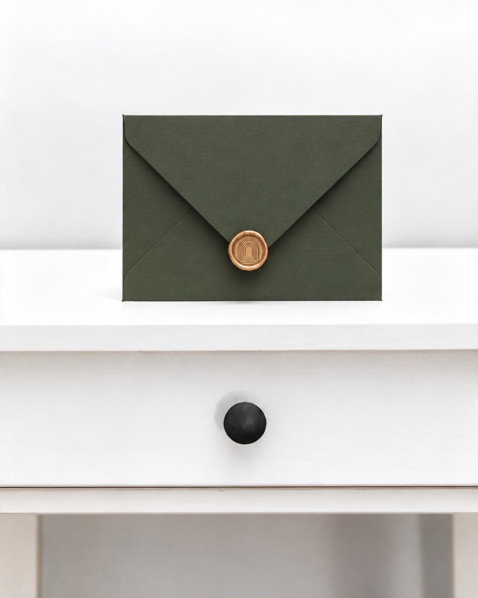 A dark green envelope closed with a gold wax seal stamped with the arch mark, standing on a white side table